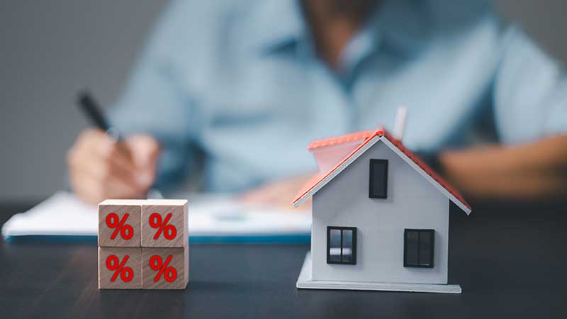 Casa e cubos de madeira com o símbolo de percentagem e um homem na retaguarda a escrever enquanto pondera a troca de casa